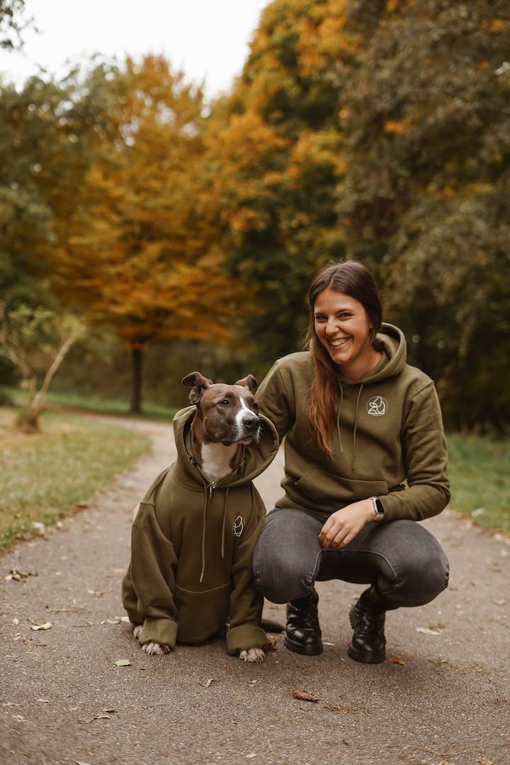 Lächelnde Person sitzt in der Hocke neben einem Hund. Beide tragen einen olivgrünen Hoodie mit dem ERBICURA Logo.